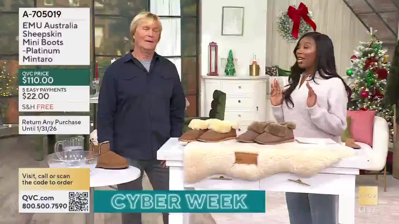 A man and a woman stand behind a table, gesturing towards several pairs of sheepskin boots. The woman, speaking animatedly, holds her hands up as if presenting the footwear on display for QVC2.
