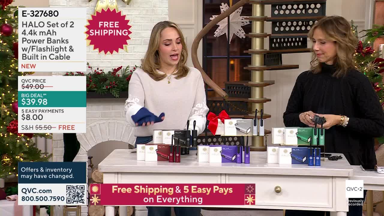 One woman holds a blue cloth, gesturing toward a table displaying colorful power banks. Her colleague, on the right, examines a black one, as the QVC2 broadcast continues.
