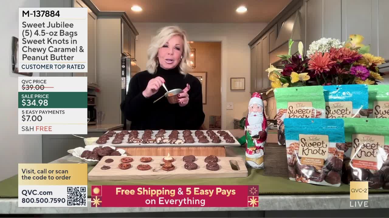 A woman on QVC2 stirs a small bowl of something brown, holding it over a tray of cookies. In the background, stacks of bags with "Sweet Knots" are visible, alongside a Santa Claus chocolate figure.
