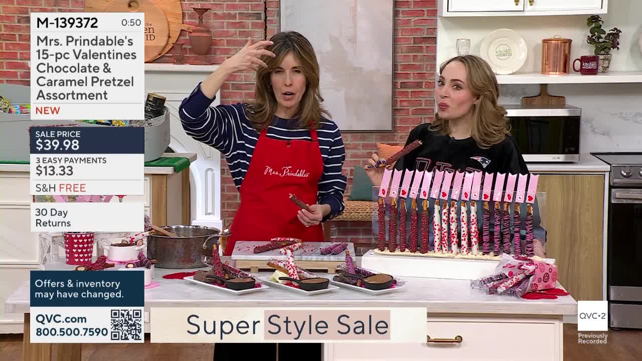 A woman in a red apron gestures with a chocolate-covered pretzel stick. Beside her, another woman holds a pretzel, a display of them lined up behind her.
