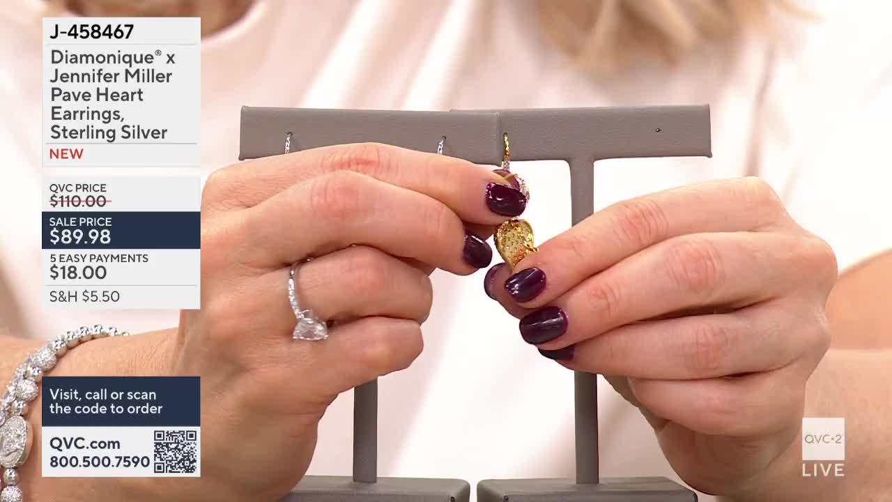 A hand with dark purple nail polish holds up a gold heart-shaped earring, its textured surface catching the light. Another hand, adorned with a sparkling ring, steadies the display.