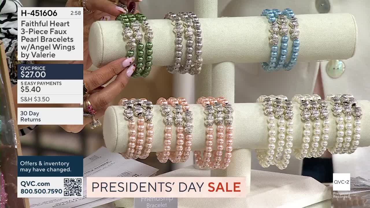 A hand reaches for a stack of green bracelets, each made of three strands of faux pearls and sparkling charms. Below, rows of pink and white pearl bracelets rest on display stands.