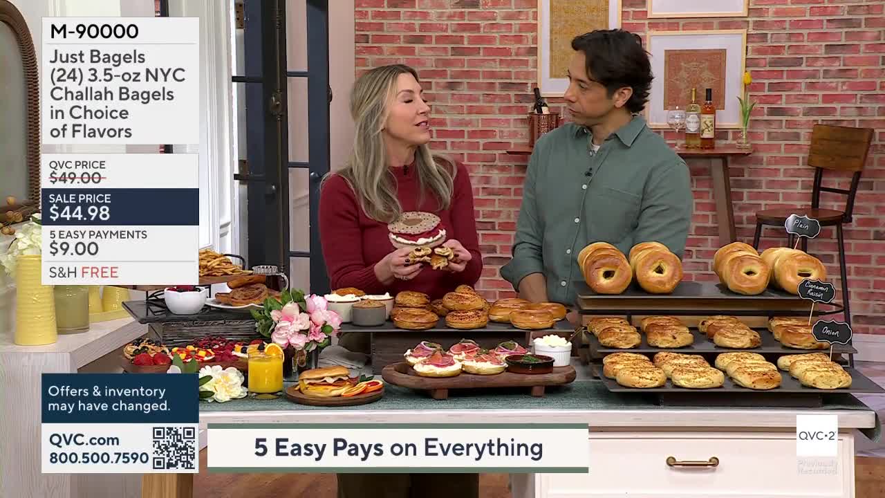 A woman holds up a bagel sandwich, explaining its features to a man. Behind them, a display showcases an assortment of bagels and pastries.