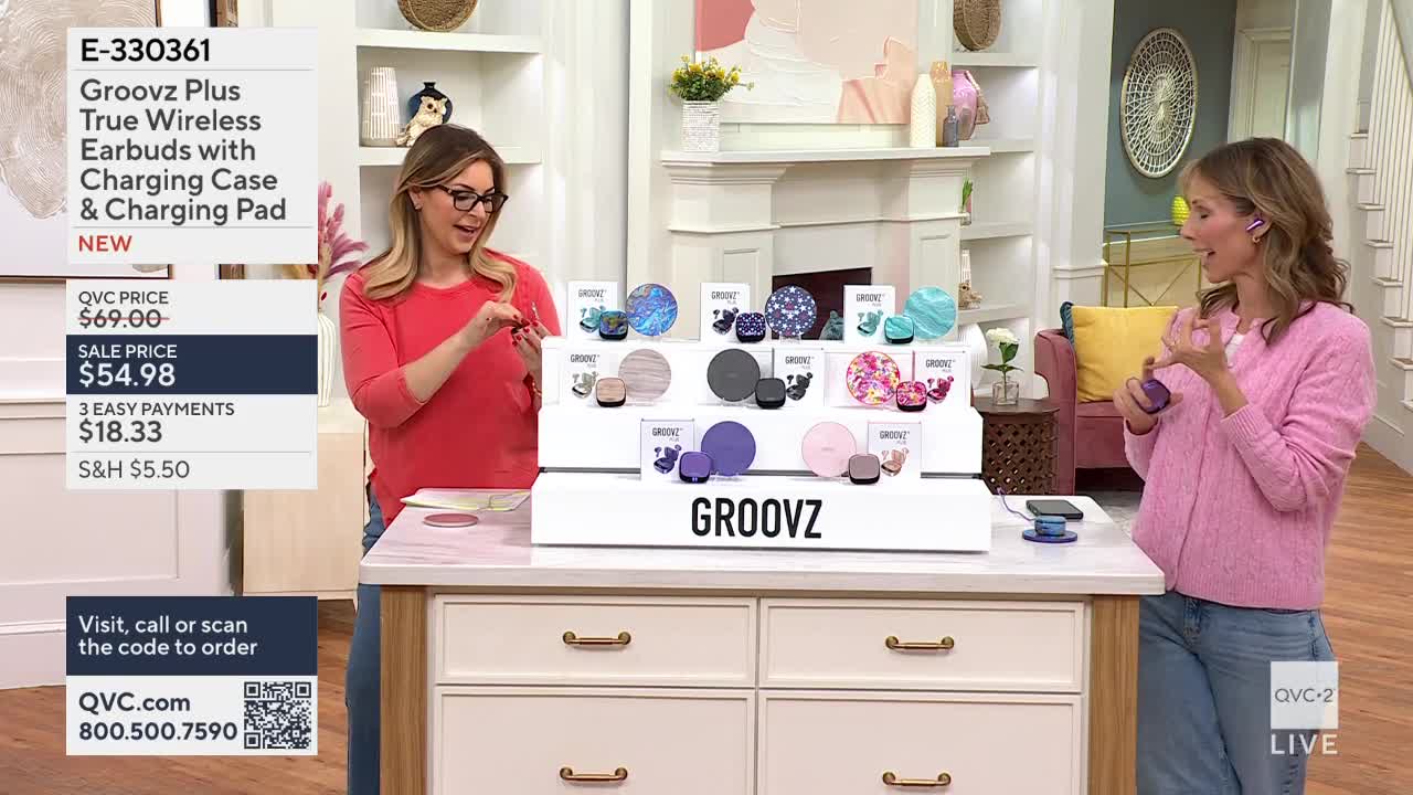 A woman in a coral shirt holds up a small red object, demonstrating it to the camera. Across from her, another woman in a pink sweater gestures with a purple earbud.