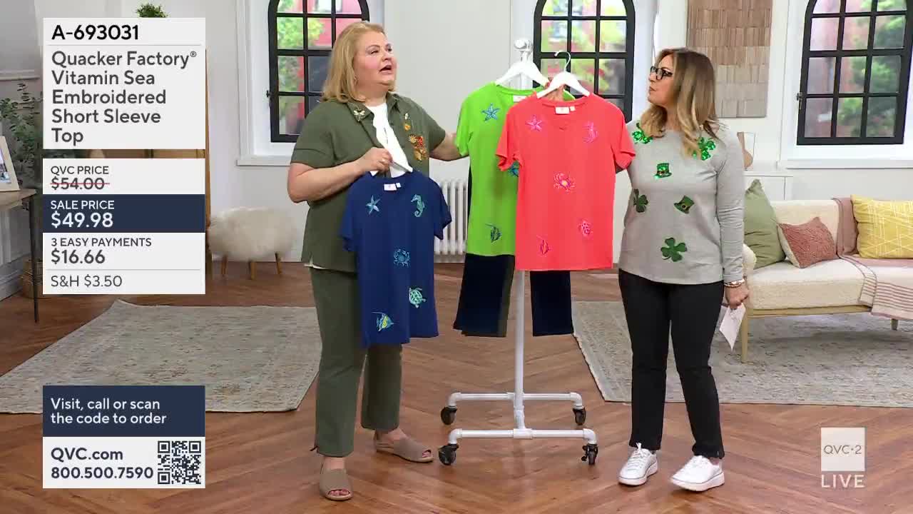 A woman on QVC2 holds up a navy blue t-shirt embroidered with starfish and seahorses. Beside her, a rack displays a bright green and a coral t-shirt, both with similar sea-themed embellishments.