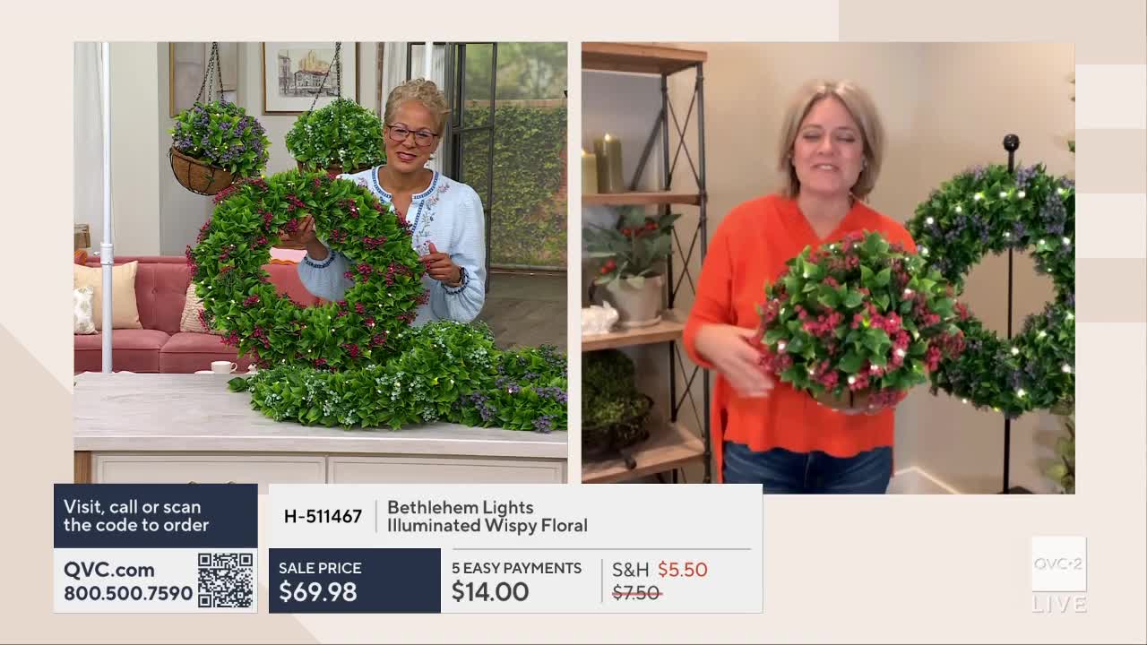 A woman in an orange shirt holds a round floral arrangement, its tiny lights twinkling. Beside her, a larger wreath, also adorned with lights, hangs on a stand.