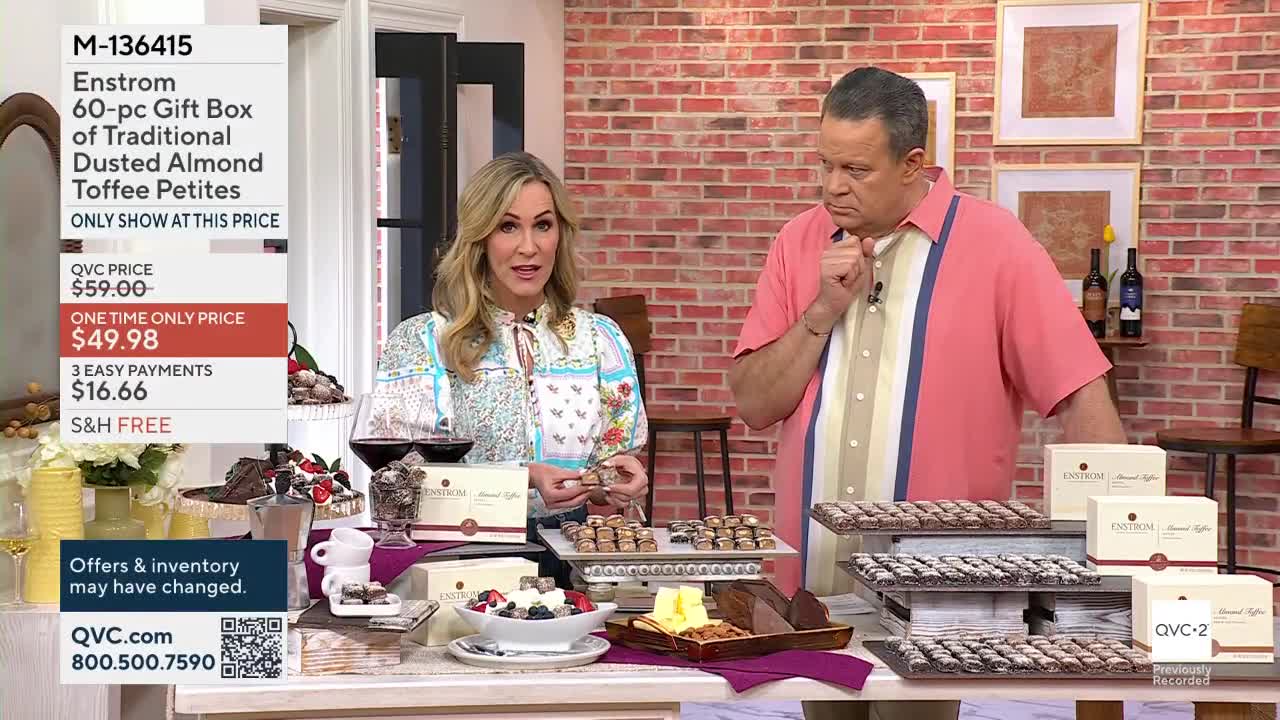 A woman holds up a small, foil-wrapped confection while a man thoughtfully strokes his chin. Rows of these chocolates are displayed on tiered stands before them, ready for purchase on QVC2.