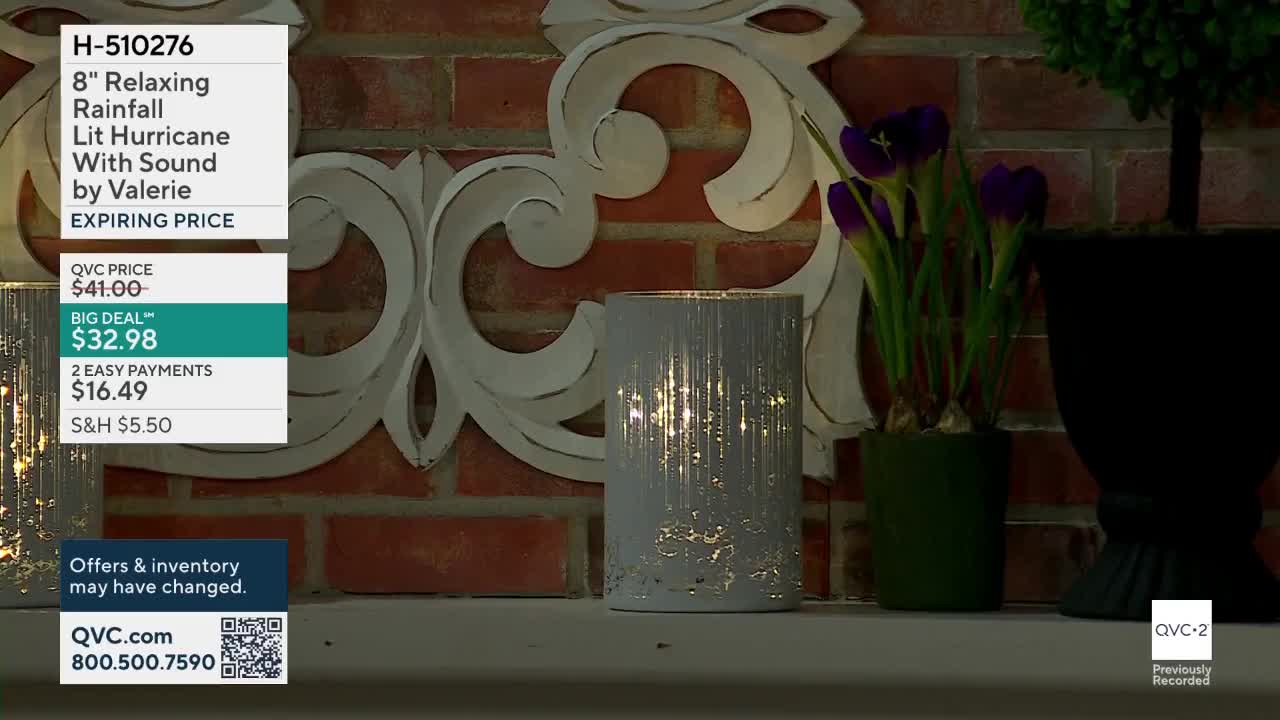 A white hurricane lantern with a textured surface glows with internal lights. Purple flowers in a green pot sit to its right, against a brick backdrop.