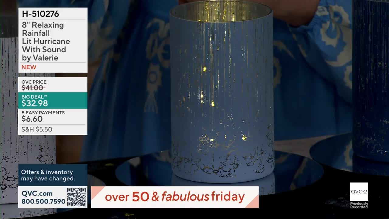 A light blue hurricane lantern with gold accents and tiny lights inside sits on a reflective surface. A blue and white patterned fabric is visible behind it.