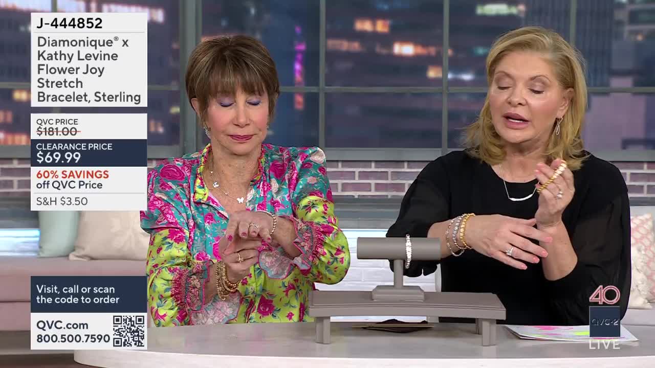 A woman in a colorful shirt adjusts a delicate bracelet on her wrist. Beside her, another woman gestures with a stack of bracelets, showcasing them to the camera.