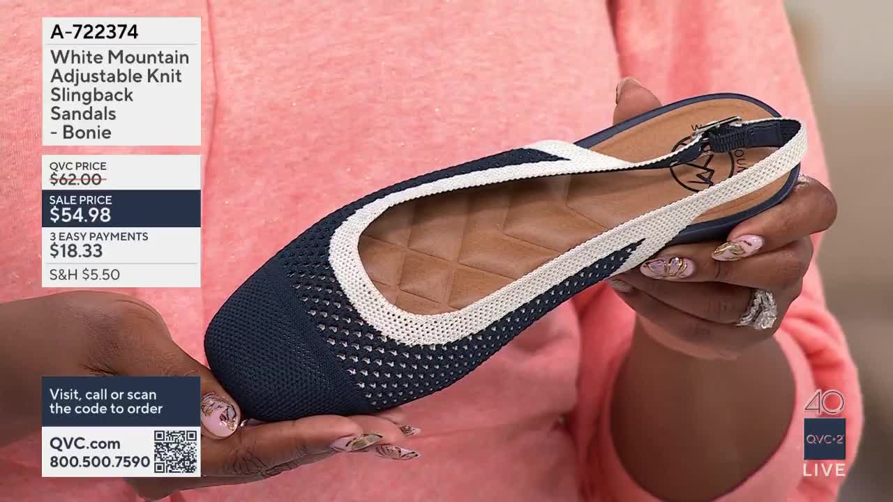 A hand turns a navy blue knit sandal with a white trim. The shoe's adjustable strap is visible, along with a quilted insole.