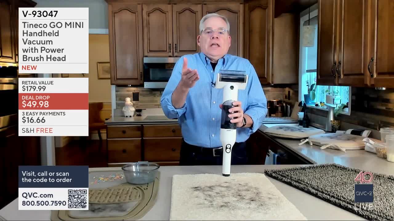 A man in a blue shirt demonstrates a white handheld vacuum on a rug. He's on QVC2, explaining the product's features.