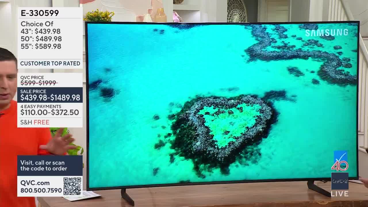 A man in an orange shirt gestures enthusiastically beside a large Samsung television displaying a vibrant aerial view of a coral reef. The screen shows pricing information for different sizes of the television, with the sale price highlighted.