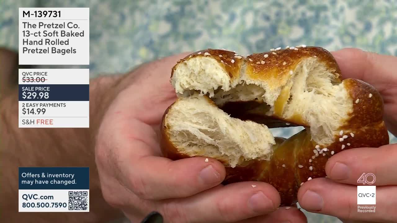 Hands pull apart a soft pretzel bagel, revealing its fluffy interior. White sesame seeds dot the golden-brown crust.