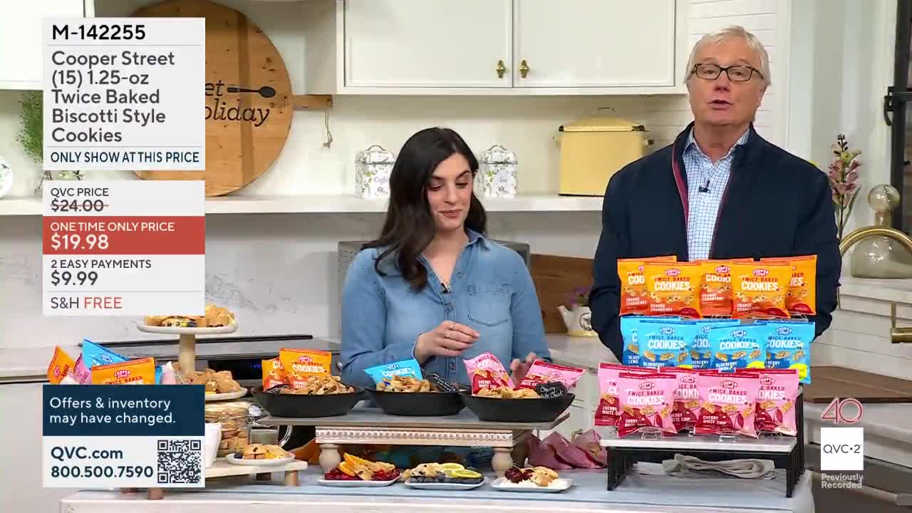A woman in a denim shirt gestures towards bowls of cookies on display. A man in a blazer holds up stacks of cookie packages, promoting them on QVC2.
