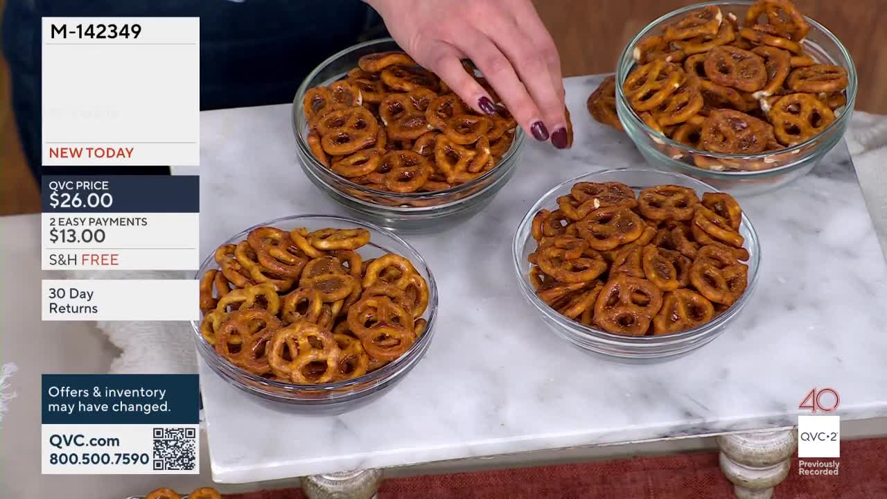 A hand with dark red nail polish reaches into a bowl of pretzels on a marble surface. Three bowls of pretzels sit on display, with a QVC2 graphic visible in the lower right corner.