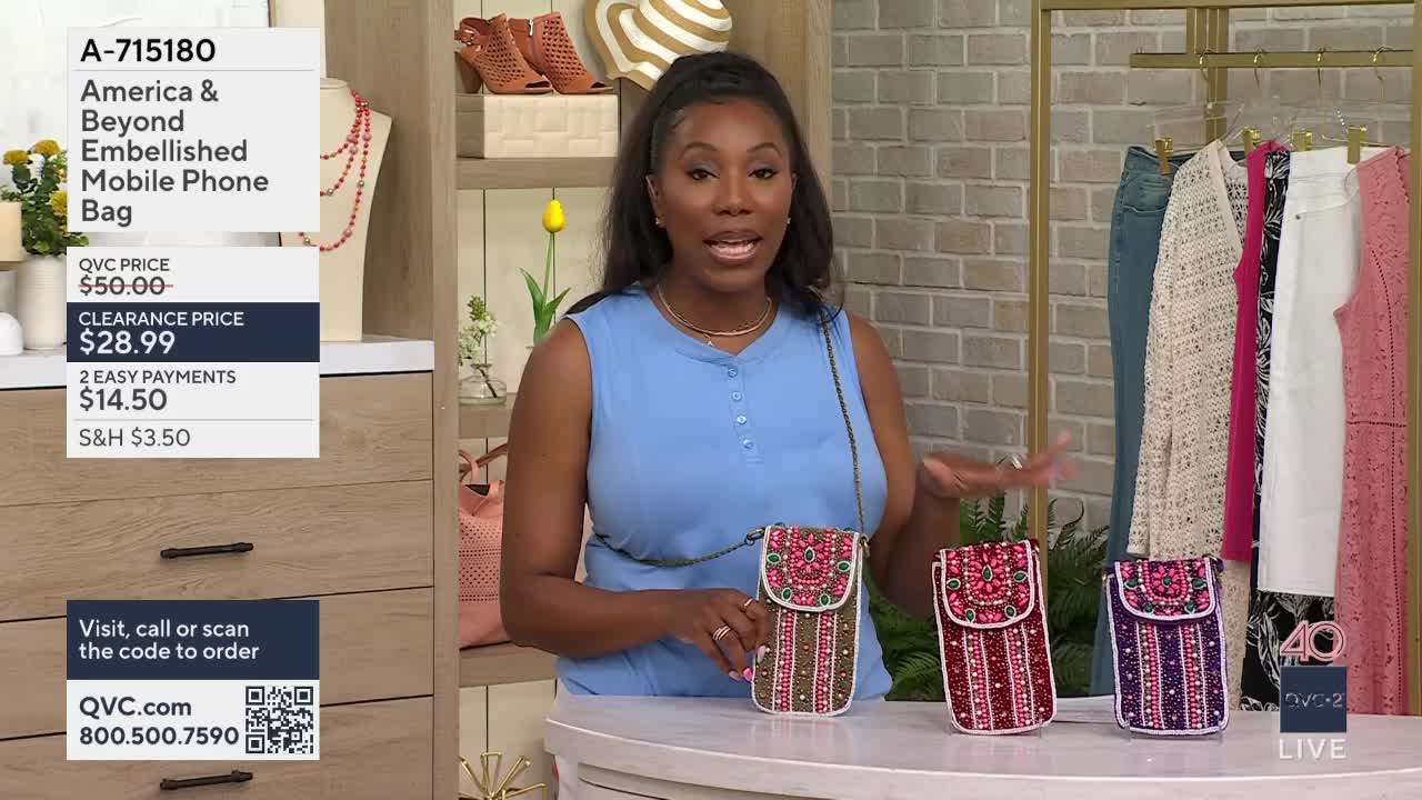 A woman on QVC2 holds up a beaded phone bag, displaying its intricate design. Three such bags, in red, pink, and purple, are lined up on a table before her.