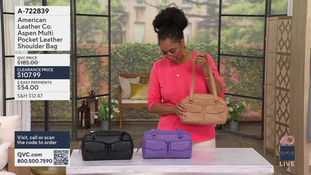 A woman in a bright pink shirt holds up a tan leather shoulder bag. In front of her, a black and a purple version of the same bag sit on a table.