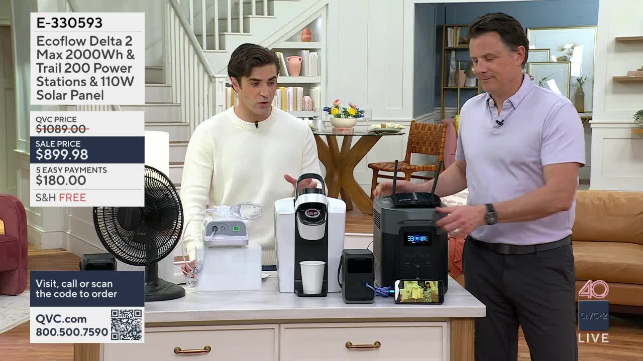 A man in a white sweater gestures toward a white coffee maker and a portable power station. Another man in a purple polo shirt stands next to the power station, his hand resting on its side.