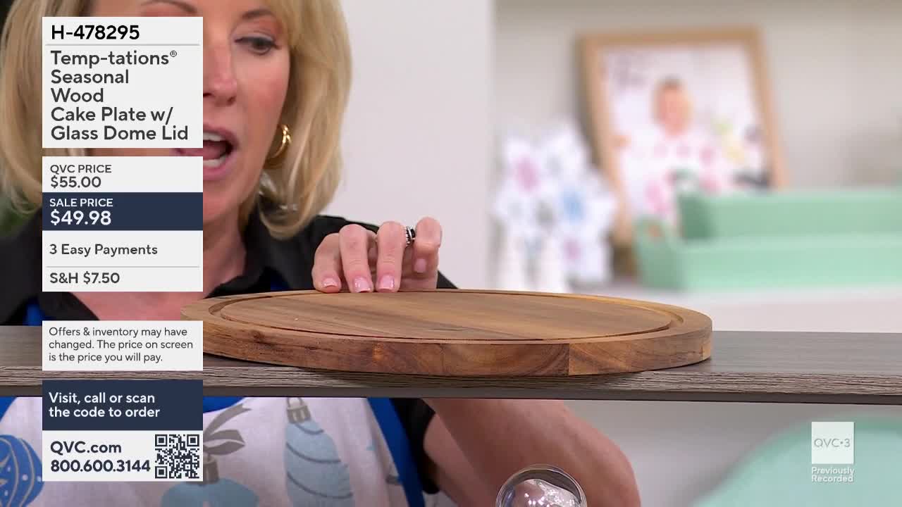 A woman on QVC3 gestures toward a wooden cake plate, showing off its details. The plate sits on a shelf, with the product information displayed on the left.
