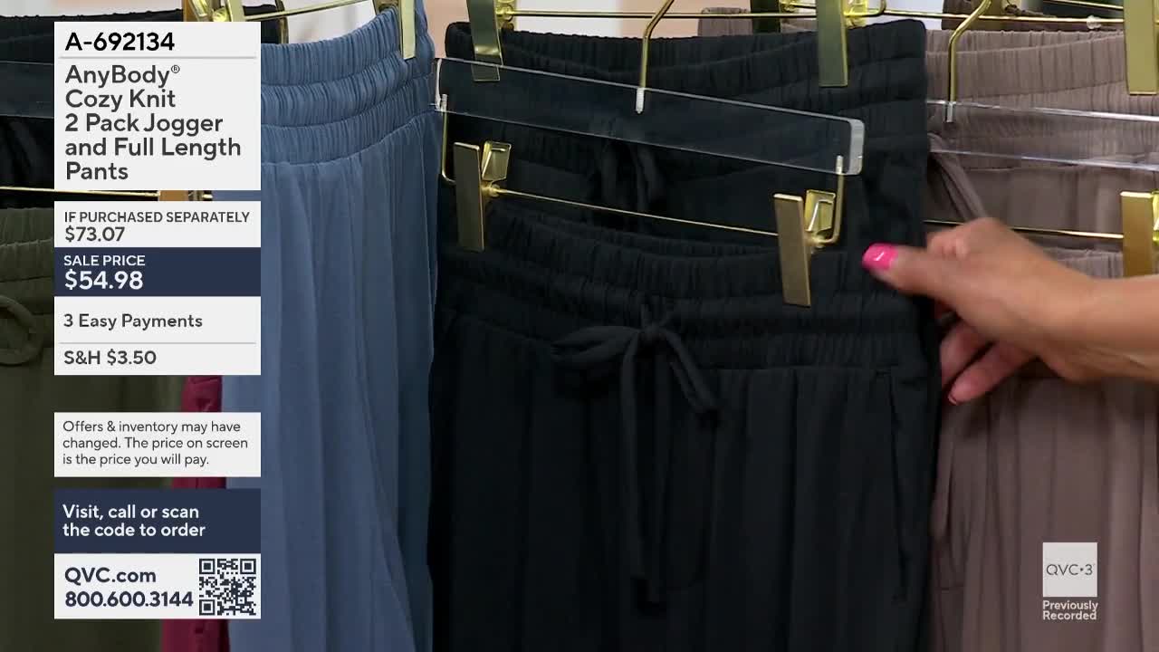 On QVC3, a hand with pink nail polish is touching the waistband of a black jogger. The pants hang from a gold hanger, next to a pair of blue joggers.
