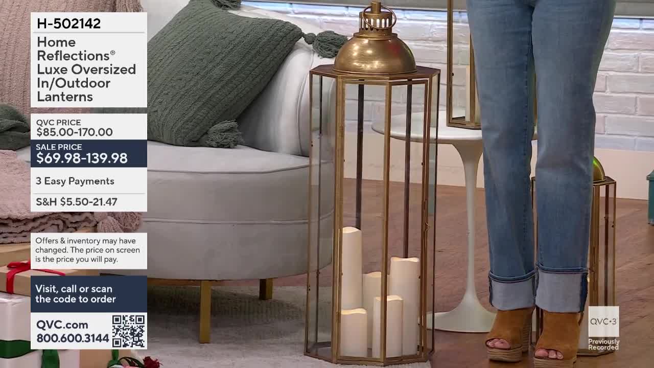 A woman in jeans and platform sandals stands near a tall, brass-framed lantern filled with white candles. The QVC3 logo is visible in the corner, and a screen displays the sale price of the featured lanterns.
