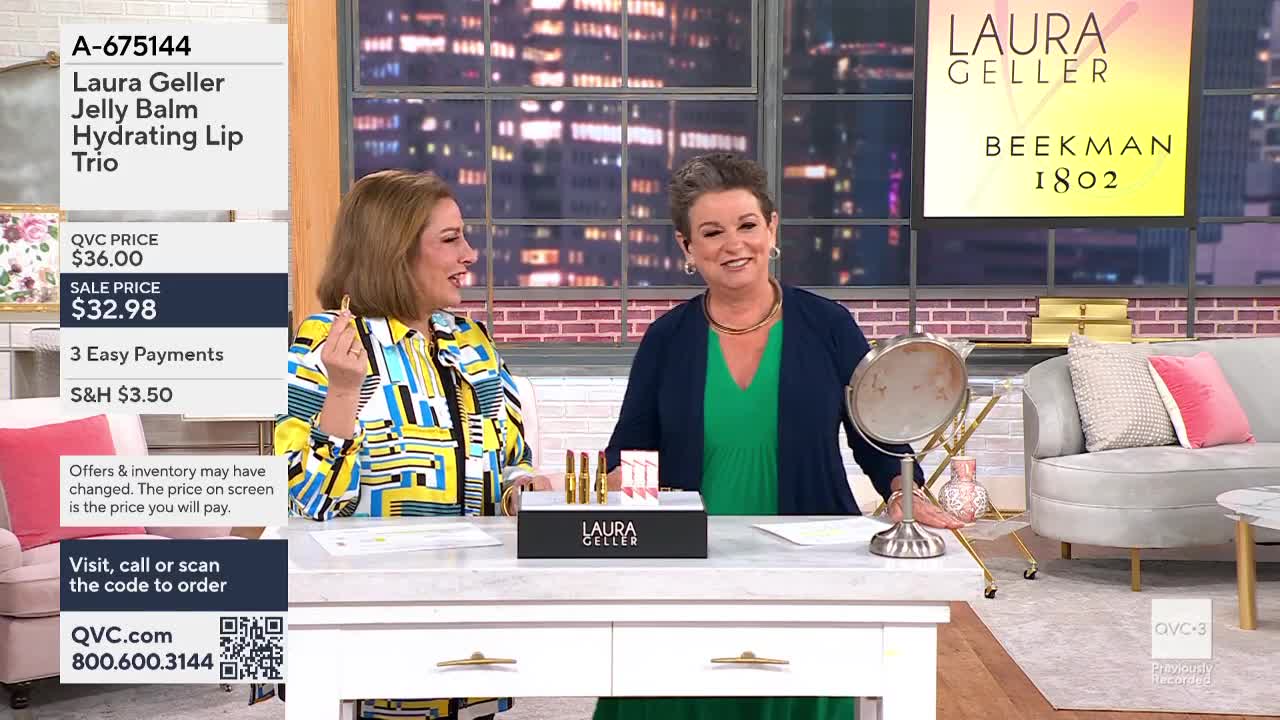 On the QVC3 set, two women are seated at a table, smiling and gesturing. One holds a lipstick, while a display of Laura Geller lip products sits between them.
