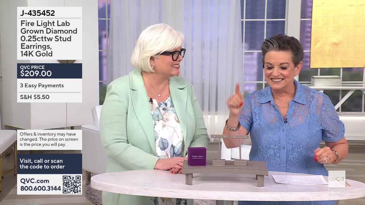 Two women on a QVC3 set are smiling, gesturing towards a small display table. One points upwards while the other looks on, seemingly engaged in a product demonstration.
