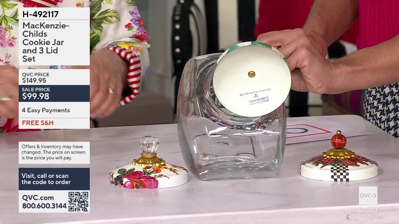 A person is holding a lid over a clear cookie jar, showing the MacKenzie-Childs logo. Two other lids with decorative tops sit on the table in front of the jar, as the QVC3 presentation continues.
