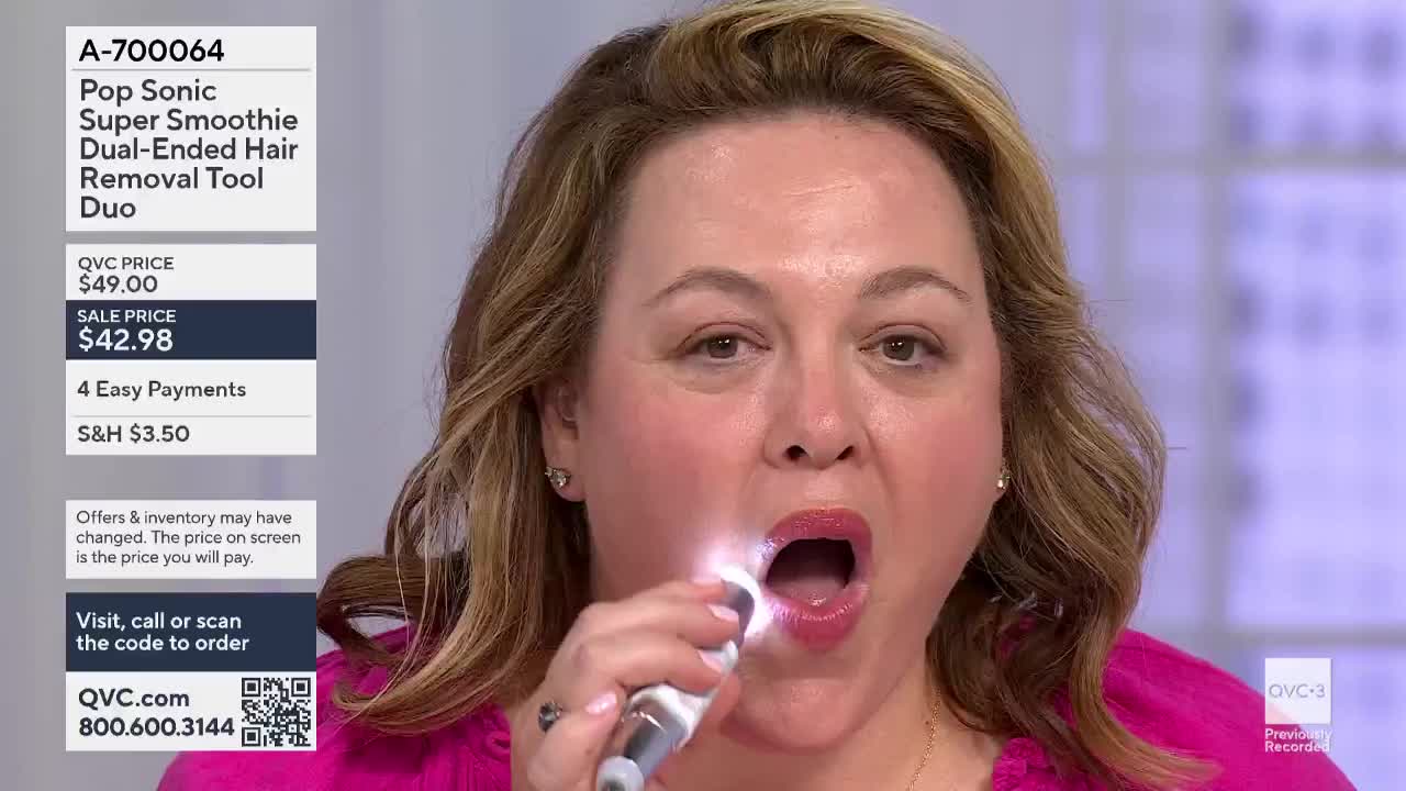 A woman on QVC3 holds a small, illuminated device up to her mouth. She's demonstrating a hair removal tool, her mouth open to show the interior.
