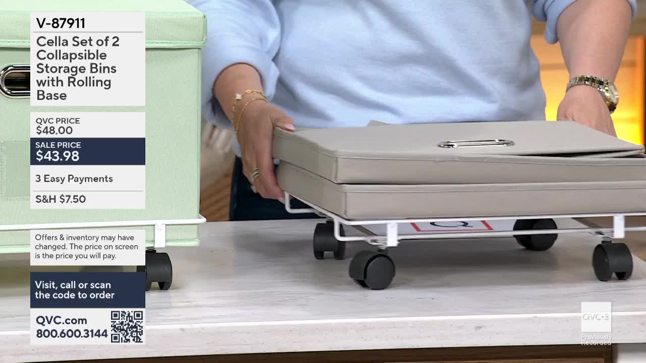 A woman on QVC3 in the United States is demonstrating a set of storage bins. She's lifting a light gray bin, which is on a rolling base, to show the product's features.
