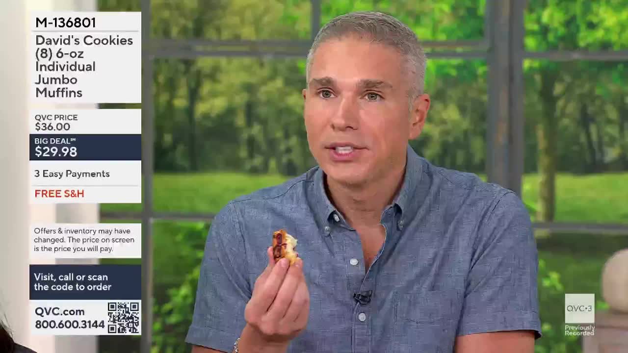 A man on QVC3 in the United States holds up a bite-sized piece of muffin, gesturing with it as he speaks. The screen displays the product details, including the price and the option to order.
