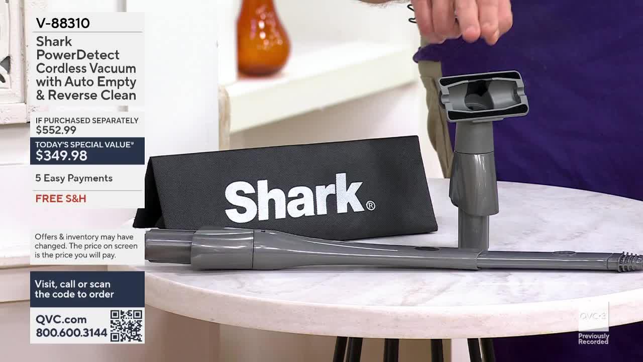 A man holds the top part of a gray vacuum, showing its features on QVC3. The vacuum's long, thin extension lies on the table next to a black bag with the Shark logo.
