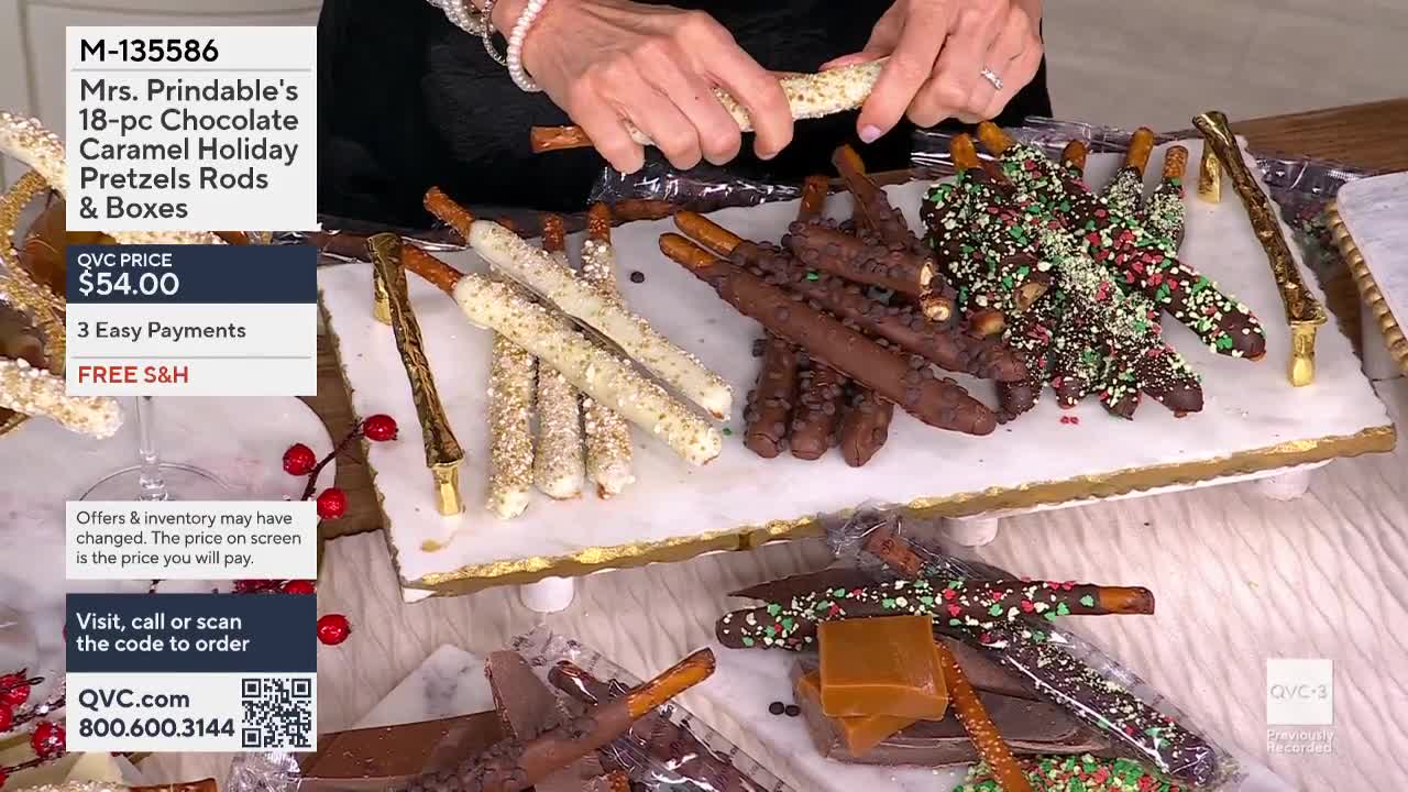 A woman on QVC3 holds a pretzel rod, coated in white chocolate and sprinkles, over a marble serving platter. The table is laden with other chocolate-covered pretzels of various types, all for sale at $54.00.
