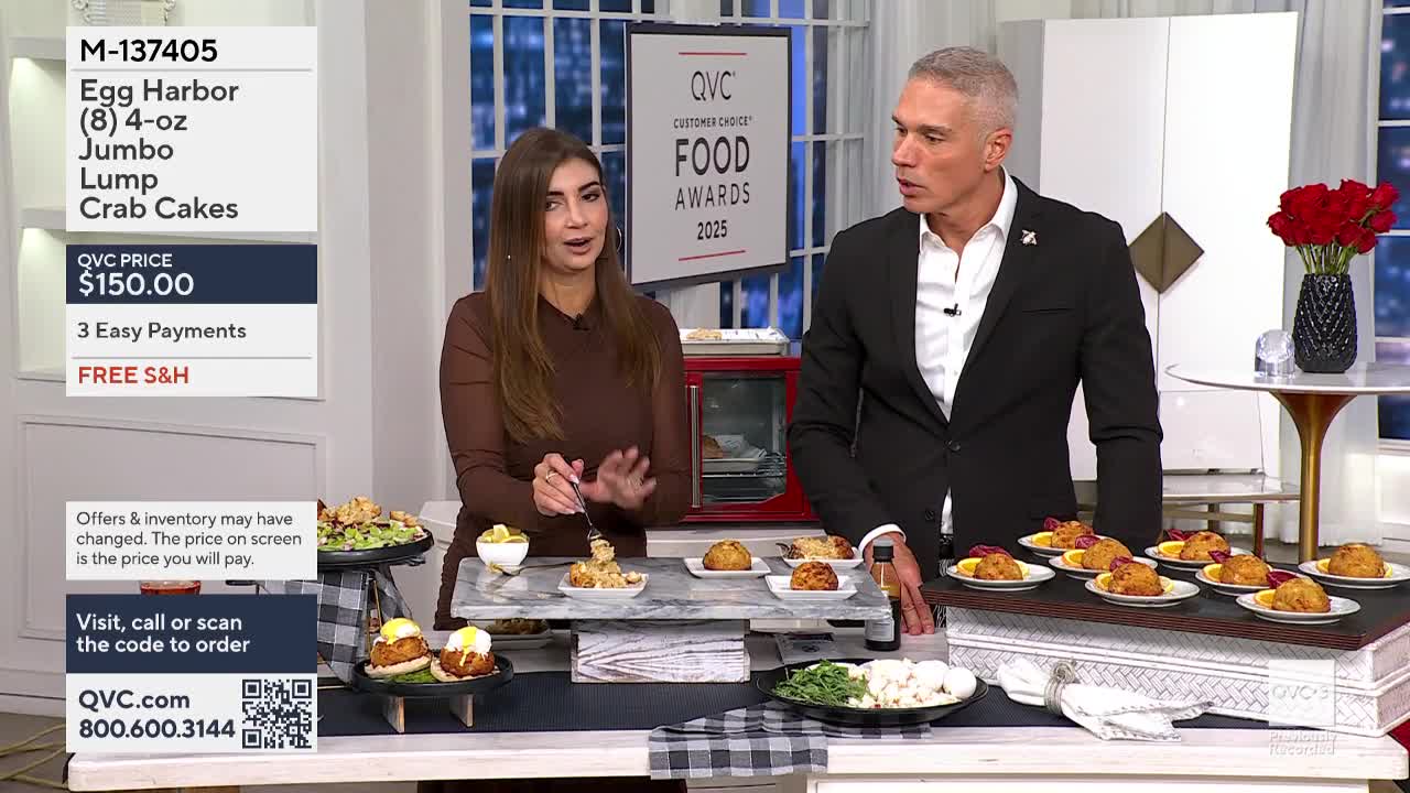 A woman in a brown dress holds a fork, gesturing toward a small plate of crab cakes. Beside her, a man in a suit stands by a table filled with plates of food, all lit for a QVC3 segment.
