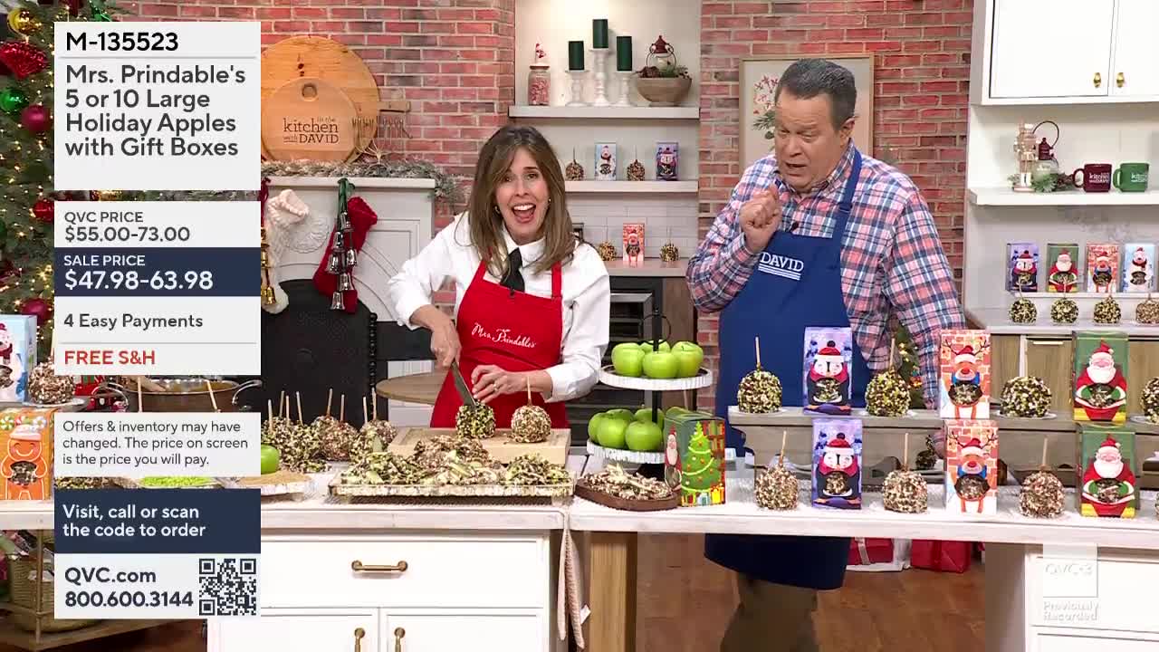 On a QVC3 set, a woman in a red apron slices a caramel apple, while a man in a blue apron gestures excitedly. Holiday-themed gift boxes and decorated apples are displayed on the counter.
