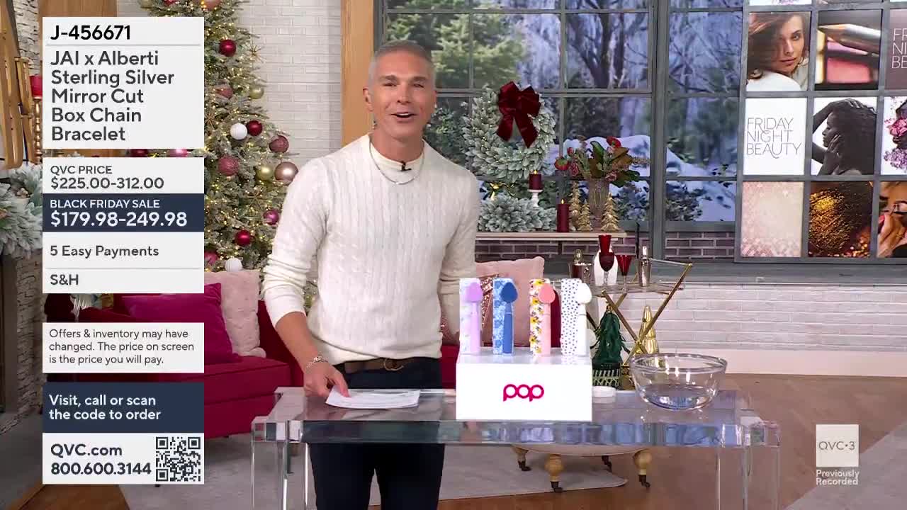 A man in a cream-colored sweater gestures towards a display of colorful items on a clear table. Behind him, a Christmas tree and a window with a winter scene set the backdrop for a QVC3 presentation.
