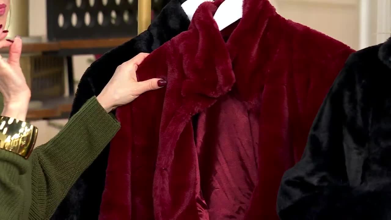 A woman's hand, with a dark red manicure, gently touches a plush, burgundy coat. Next to it, a black coat hangs on a white hanger, both on display for a QVC3 presentation.
