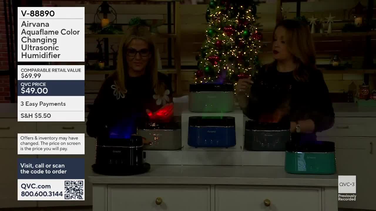 Two women stand behind a display of small humidifiers, each emitting a colored, flickering light. They're on QVC3, and one woman gestures toward the products as the other smiles and looks at the camera.
