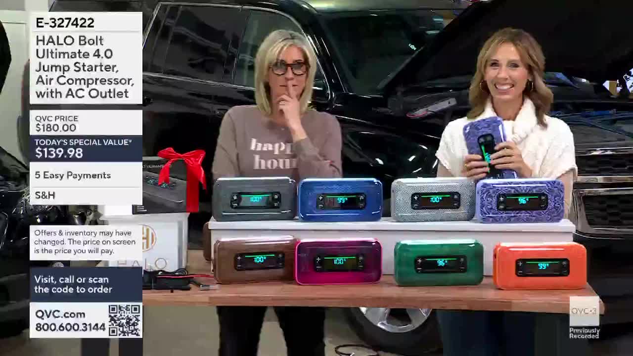Two women stand behind a table, showcasing colorful jump starters on QVC3. One woman holds a matching case, while the other gestures towards the products.
