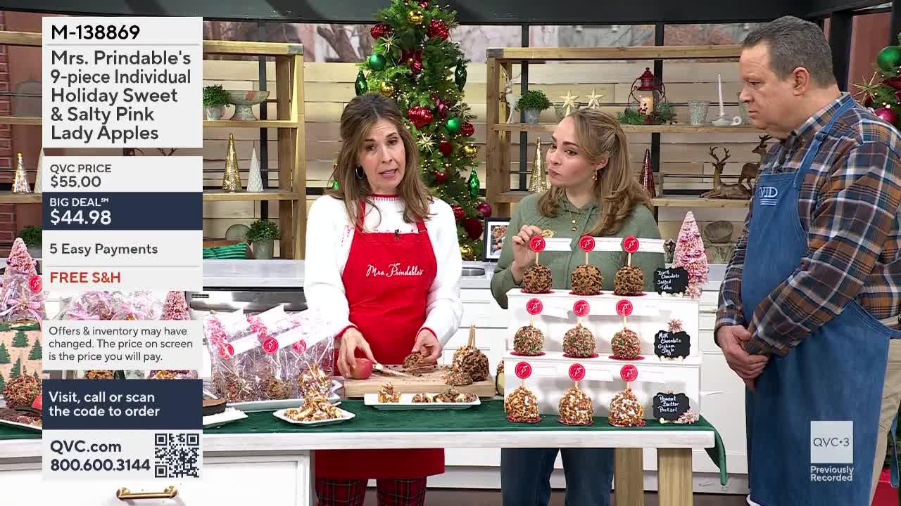 On the QVC3 set, a woman in a red apron holds a pink apple, while two other people look on. Rows of caramel apples sit on display behind them, ready for sale.
