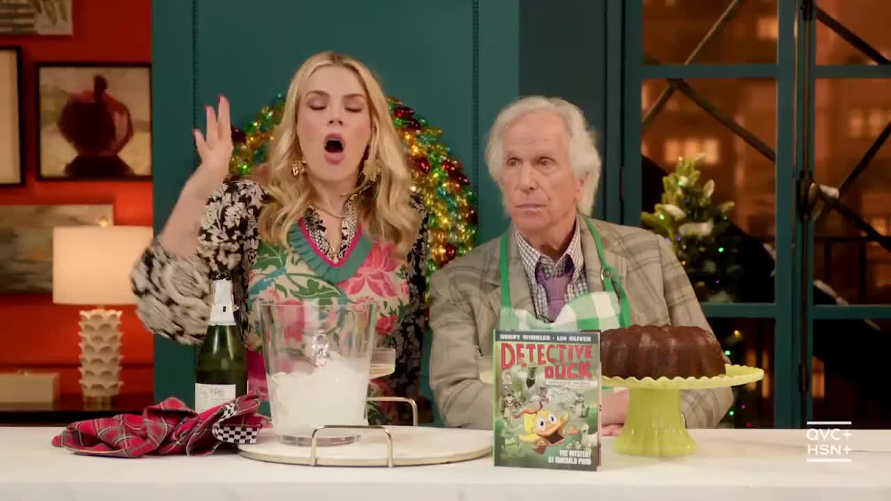 A woman in a colorful top gestures wildly, her mouth open as if mid-sentence. Beside her, a man in a plaid jacket and apron looks on, seemingly unfazed, as they appear to be on QVC3.
