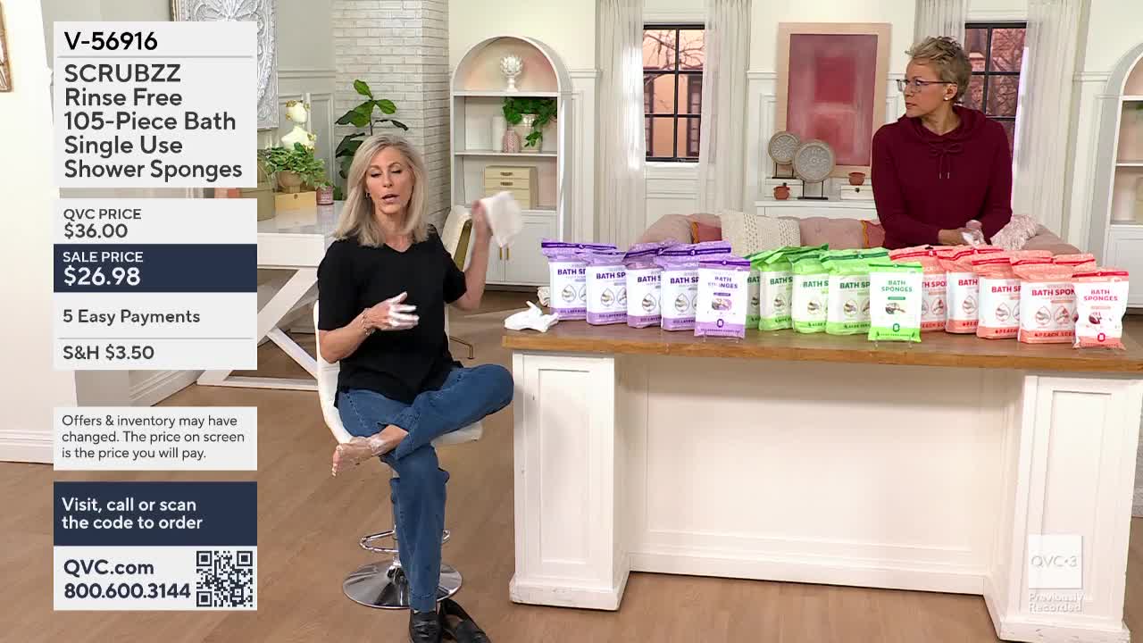 A woman in jeans and a black top holds up a white sponge, gesturing with her other hand. Rows of packaged bath sponges line a wooden counter behind her.