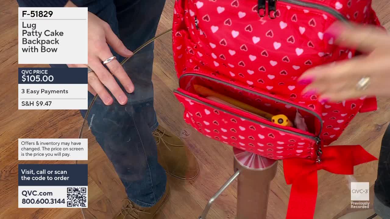 A hand with bright pink nails reaches into the open pocket of a red backpack covered in white hearts. The backpack is secured to a stand with a large red bow.