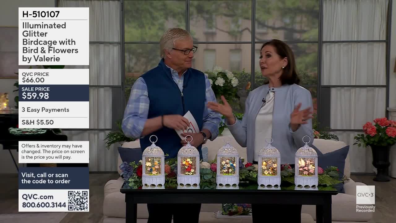 A man and a woman stand behind a table displaying four illuminated birdcages. The birdcages, each with a delicate white metal frame, contain colorful floral arrangements and tiny birds. The woman gestures enthusiastically as she speaks, while the man listens attentively, holding a brochure.