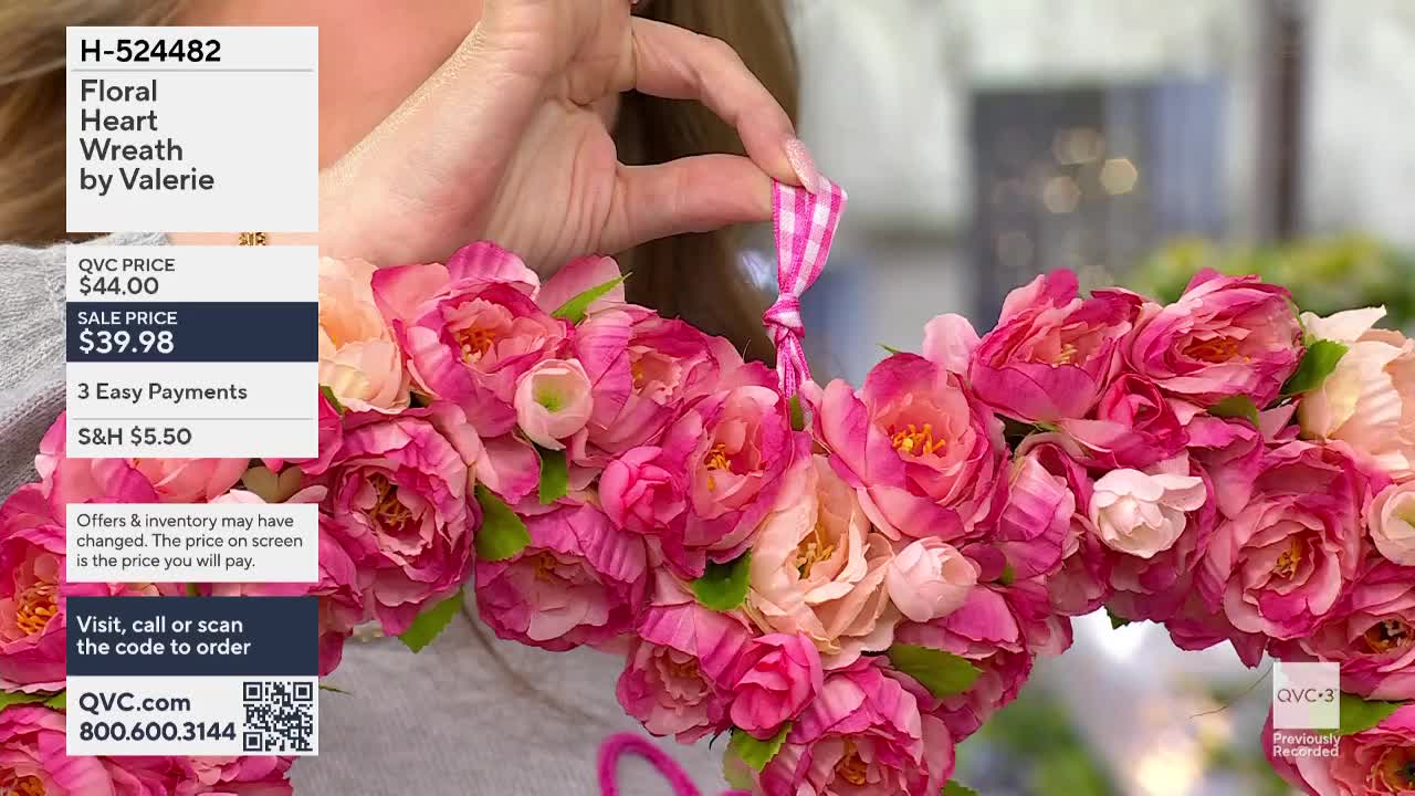 A hand holds a pink gingham ribbon tied to a heart-shaped wreath overflowing with vibrant pink and peach blossoms. This QVC item, the Floral Heart Wreath by Valerie, is currently on sale for $39.98.