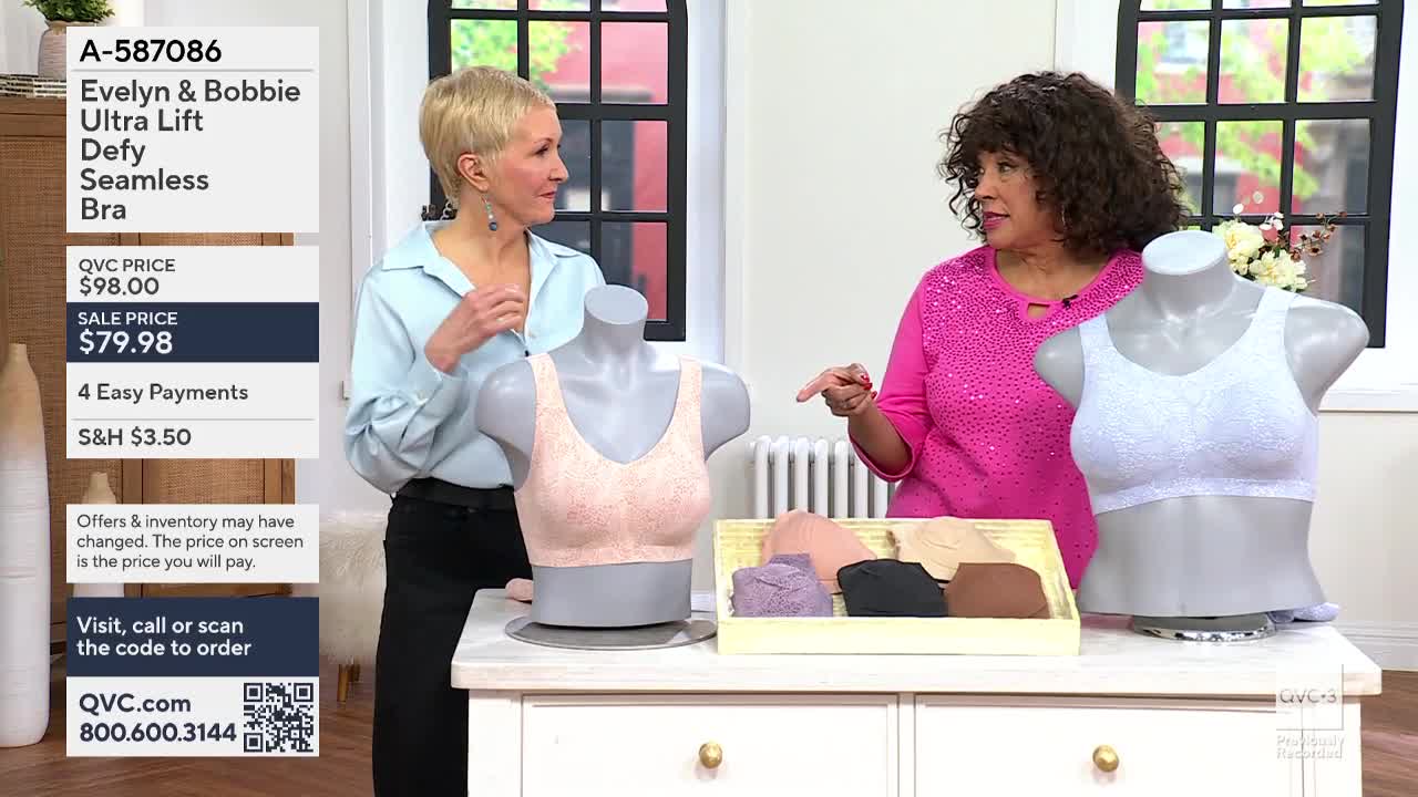 A woman in a bright pink shirt points to a white bra on a mannequin. Beside her, another woman adjusts the strap of a peach-colored bra on a different mannequin. A display tray holds several more bras in various neutral shades.