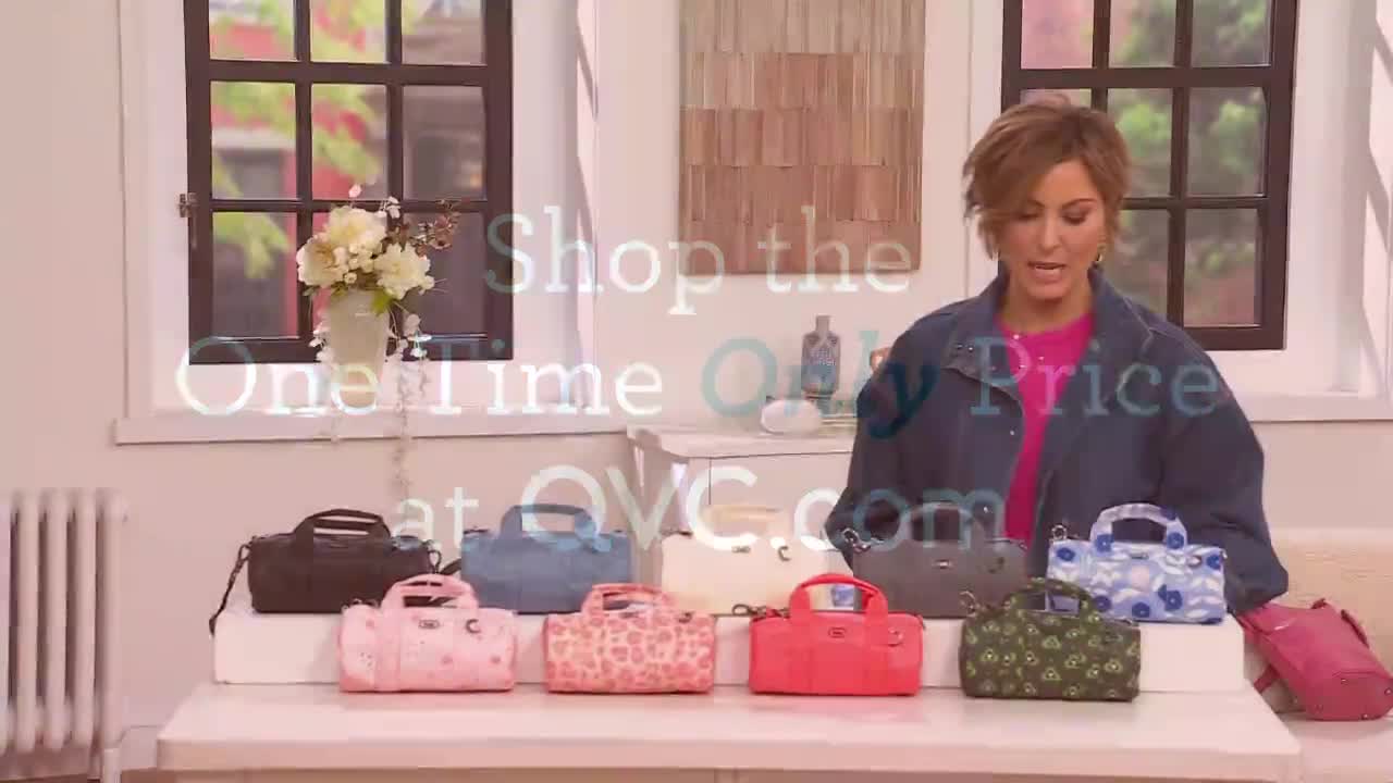 A woman on QVC is showing off a collection of colorful handbags. The bags are displayed on a table in front of her, with a window and a vase of flowers in the background.