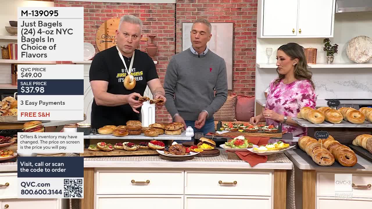 A man holds up a bagel, gesturing with his other hand as he speaks. A second man stands beside him, looking on, while a woman to the right arranges food on a display.