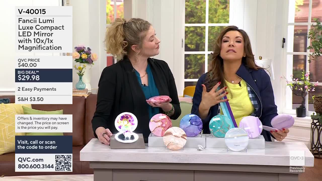 Two women are on a set, presenting several compact mirrors. One woman holds a pink mirror while gesturing, and the other touches her chest, looking up.